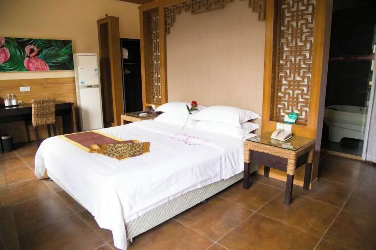A hotel room with a bed, a bedside table with a phone, a decorative screen, a desk with a chair, and a painting on the wall.