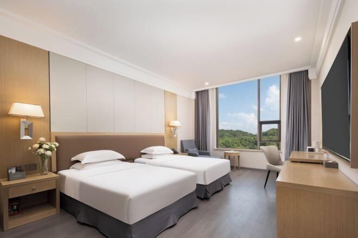 A hotel room with two beds, a desk, a chair, a small table, and a large window with a view of greenery.