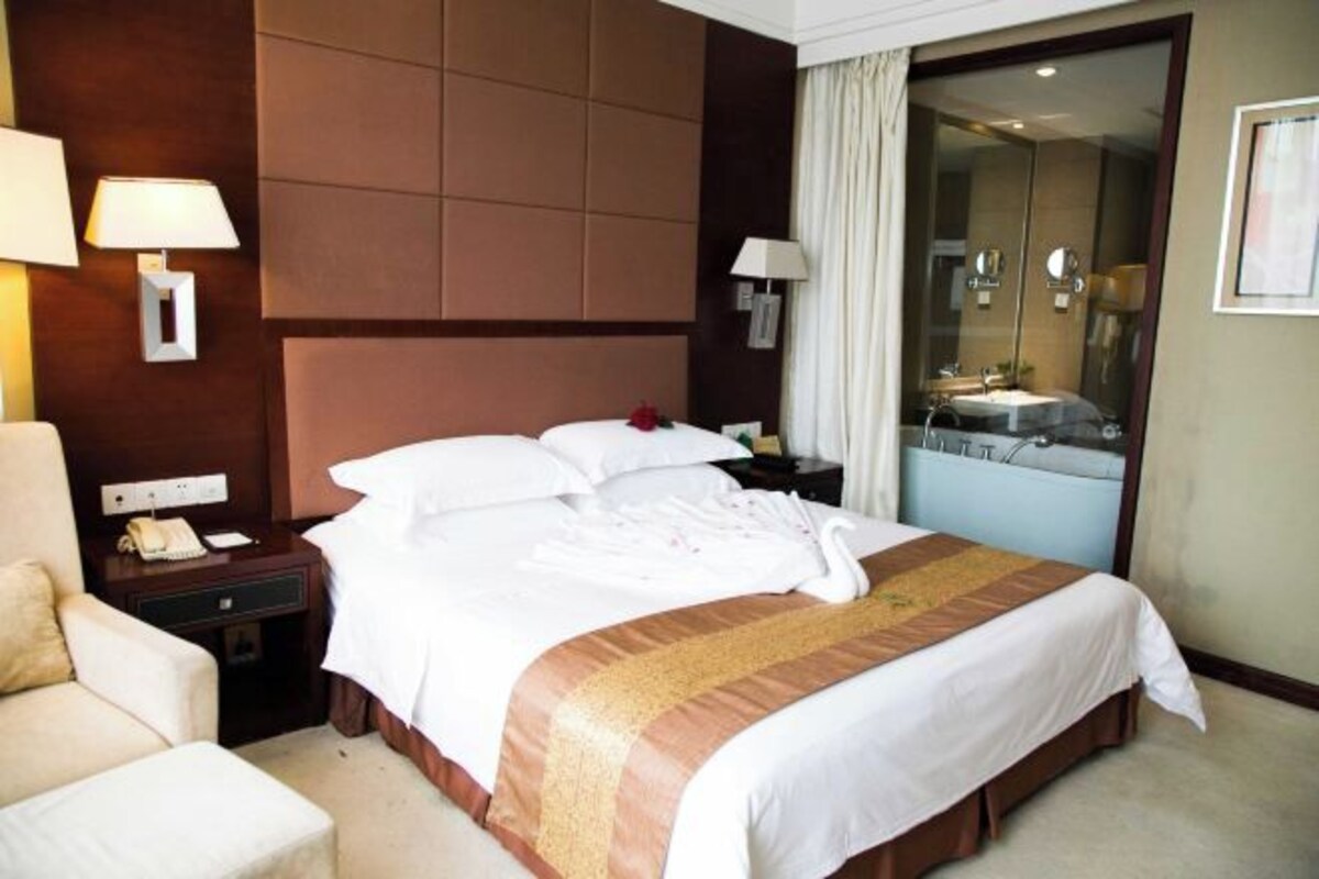 A hotel room with a large bed, a nightstand with a lamp, a chair, and a bathroom visible through a glass door.