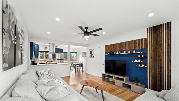 Een moderne woonkamer met een grijze bank, een houten salontafel en een televisie. Achtergrond toont een keuken met blauwe kastjes en een eethoek.