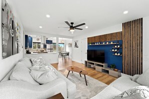 Un salon moderne avec un canapé gris, une table basse en bois et un téléviseur. On aperçoit une cuisine à l’arrière-plan, avec des meubles bleus et un coin repas.