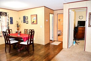 A dining area with a wooden table and chairs, a red tablecloth, and a vase with flowers. There are framed pictures on the wall and a door leading to another room.