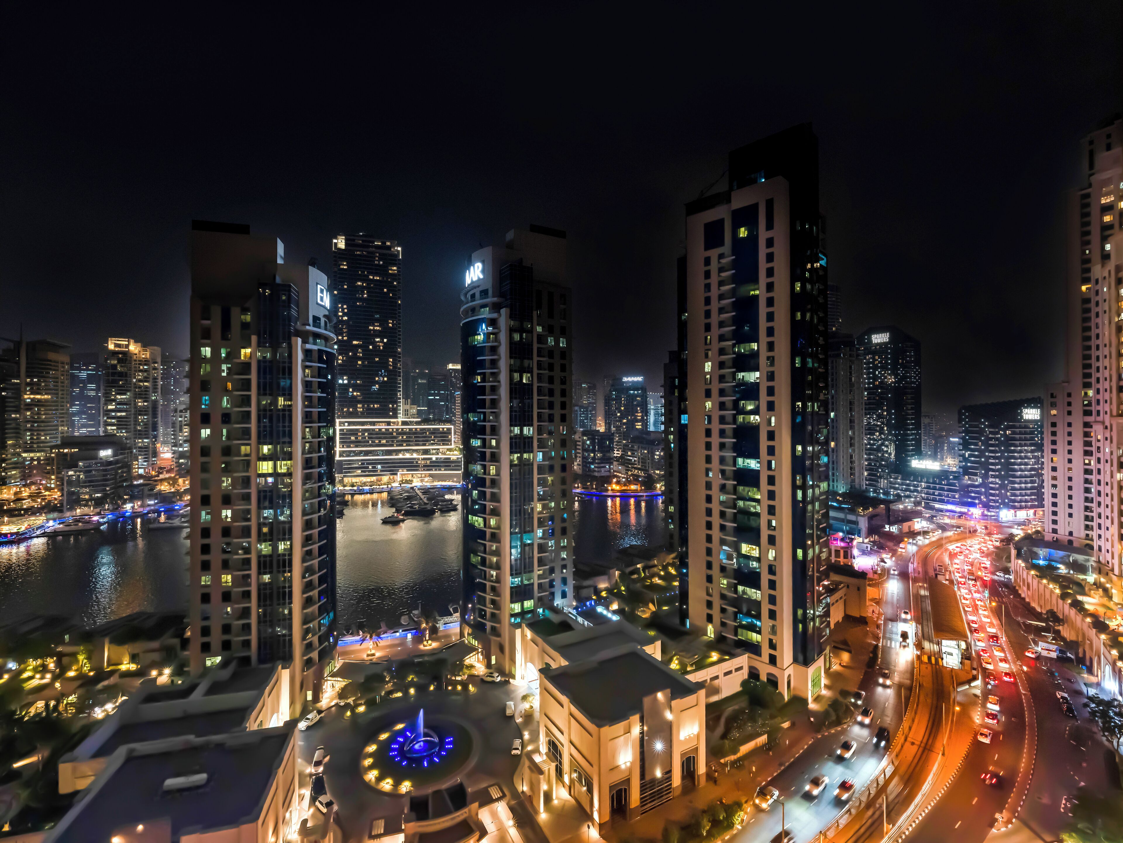 A nighttime cityscape with high-rise buildings, a waterfront, and a roundabout with a central fountain.
