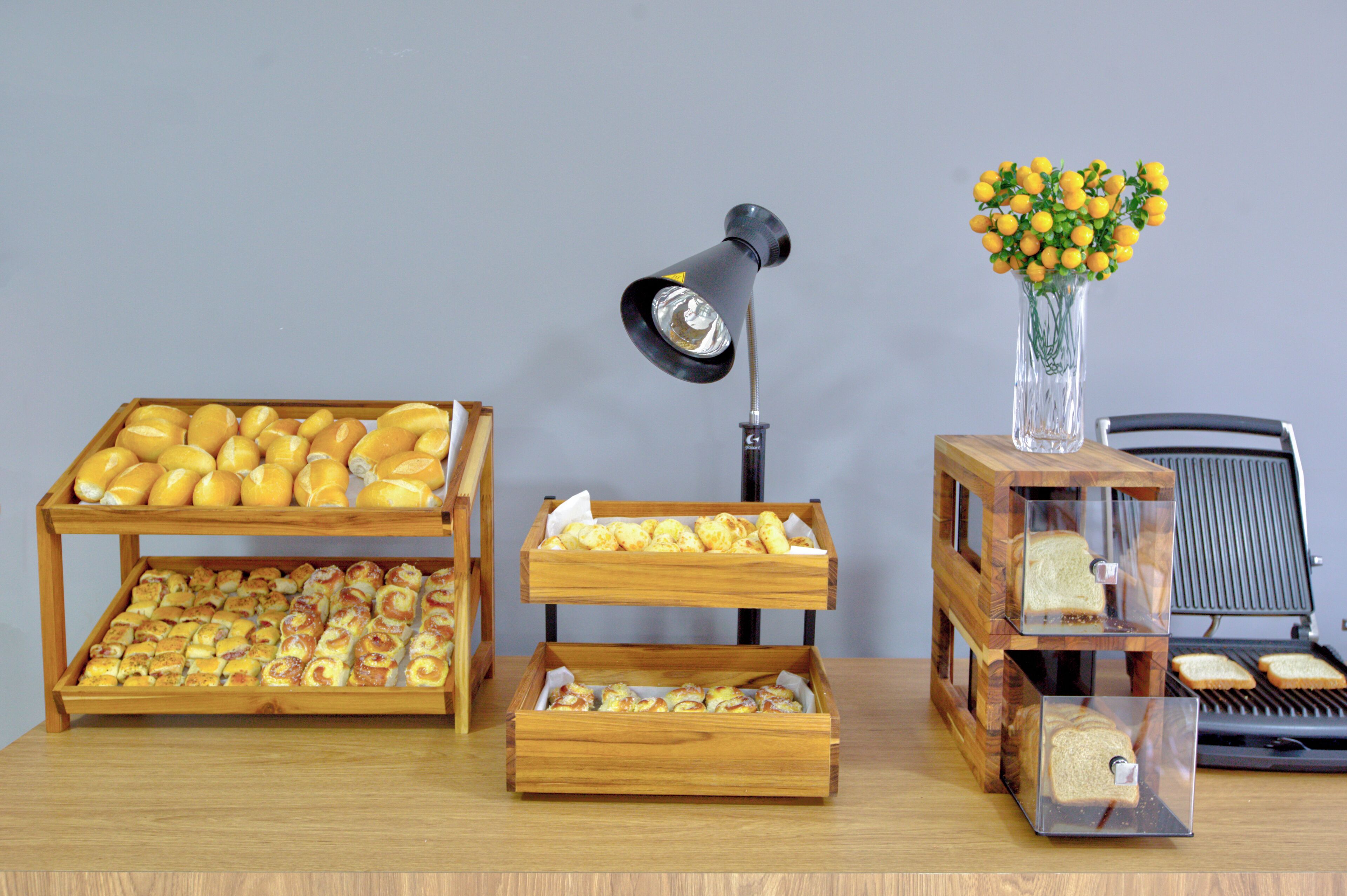 A wooden table with trays of pastries and bread, a lamp, and a vase with yellow flowers.