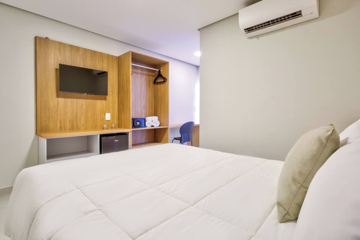 A hotel room with a bed, a desk, a chair, a wardrobe, and an air conditioning unit.