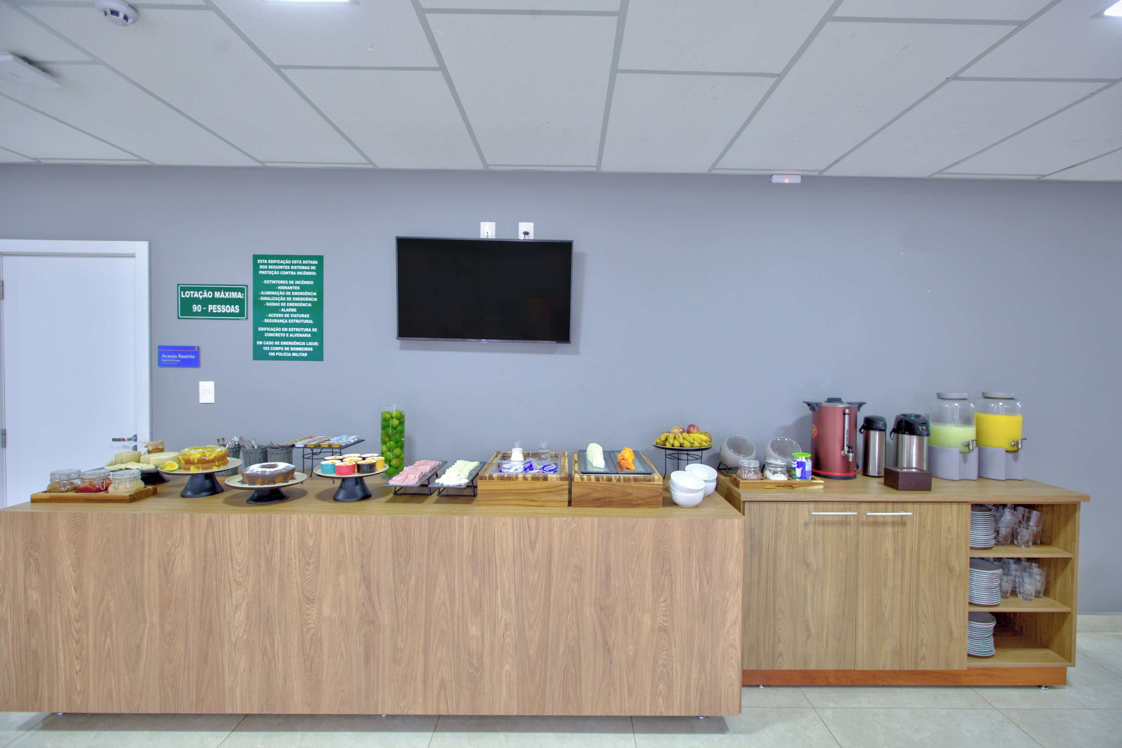 A buffet area with a variety of food items and beverages, a television mounted on the wall, and a sign indicating a maximum capacity of 90 people.