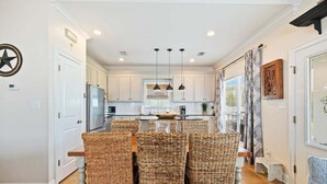 A modern kitchen with white cabinetry, a central island, and a dining area with wicker chairs. - Bear Point 3BR House Orange Beach Bring Your Boat! (Orange Beach)