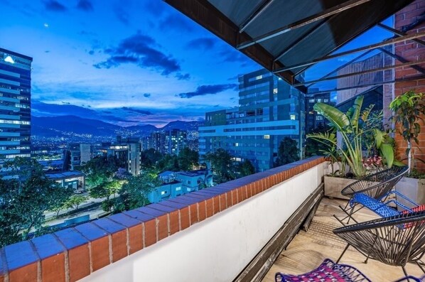 A balcony with a city view at dusk, featuring a brick wall, a glass railing, and two chairs.