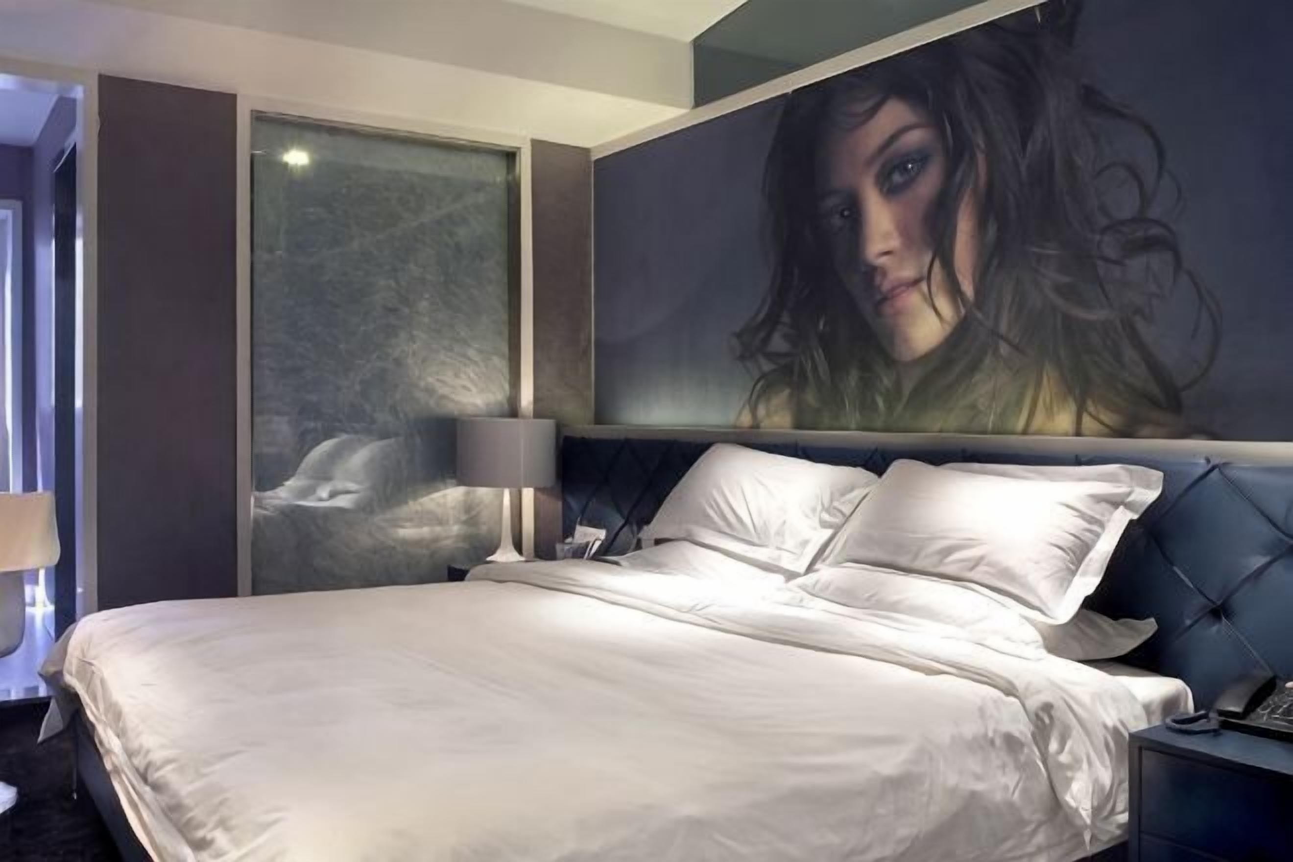 A modern hotel room with a large bed, a nightstand, a lamp, and a painting on the wall.