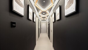 A hallway with framed pictures on the walls, a lighted ceiling, and a door at the end.