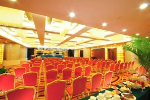 Un salón de banquetes con filas de sillas rojas, una larga mesa con comida y bebidas, y un escenario al frente.