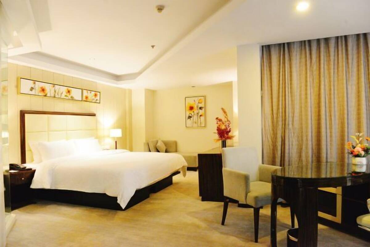 A hotel room with a bed, bedside table, lamp, chair, and a dining table with a vase of flowers.