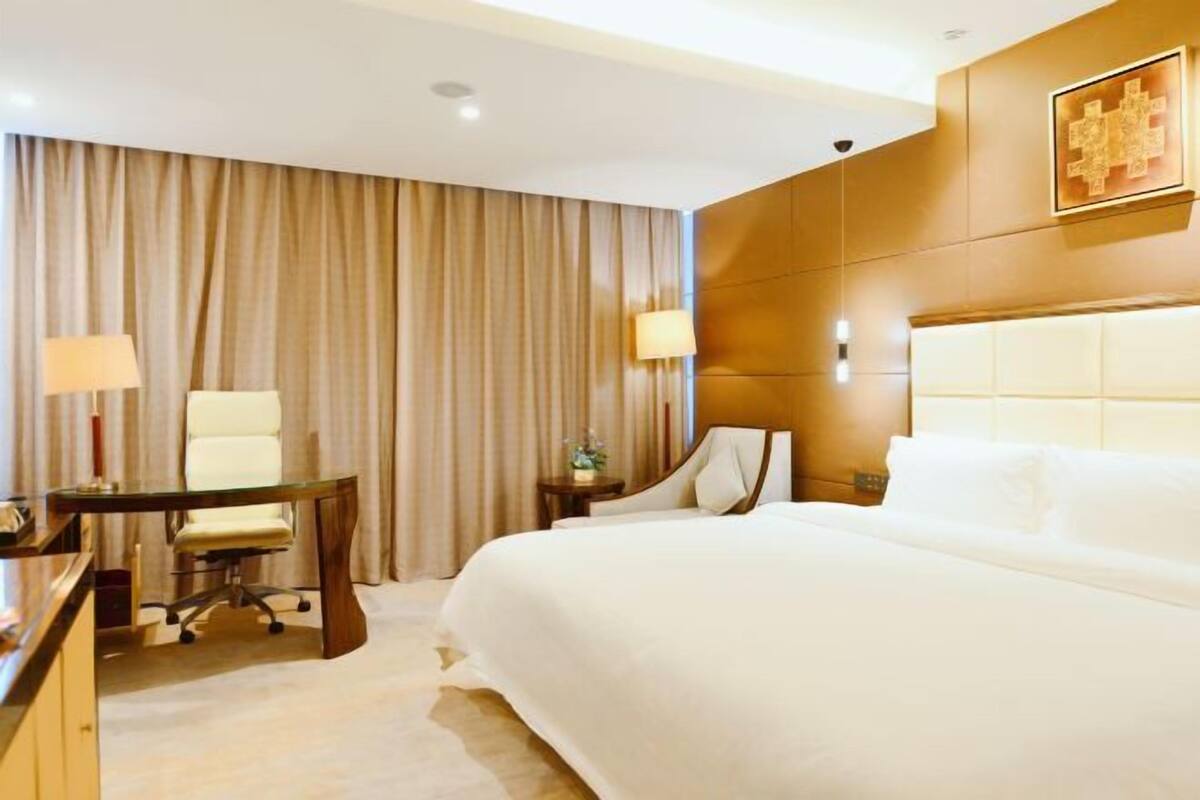 A hotel room with a large bed, a desk, a chair, a lamp, and a wall-mounted artwork.