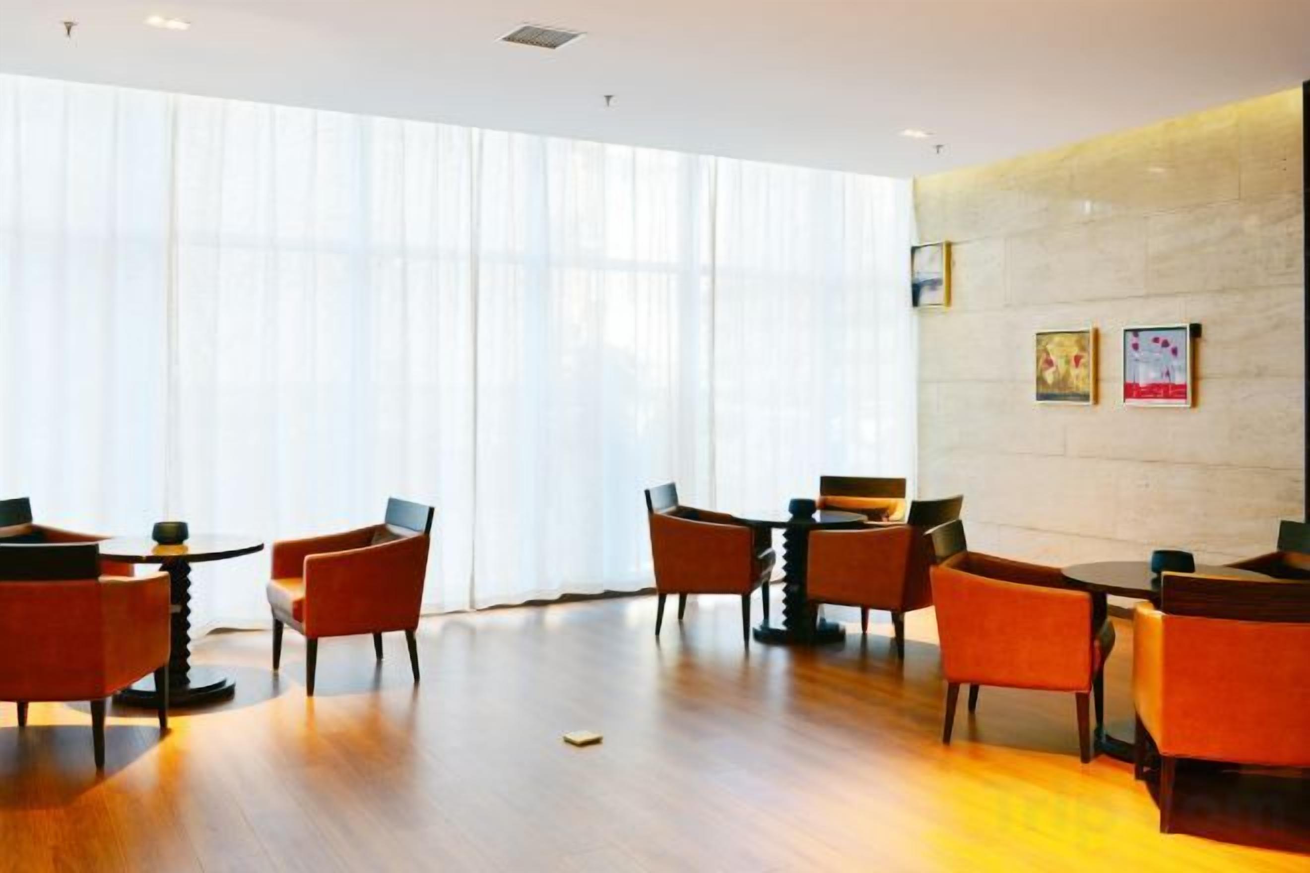 A hotel lobby with a wooden floor, orange chairs, and a large window with sheer curtains.