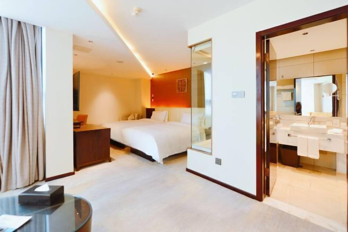 A hotel room with a bed, a desk, a glass partition, and a bathroom with a mirror and sink.