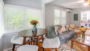 Ein Wohnzimmer mit einem runden Holztisch, zwei grauen Sesseln, einem grauen Sofa mit dekorativen Kissen und einem Couchtisch mit Blumen.