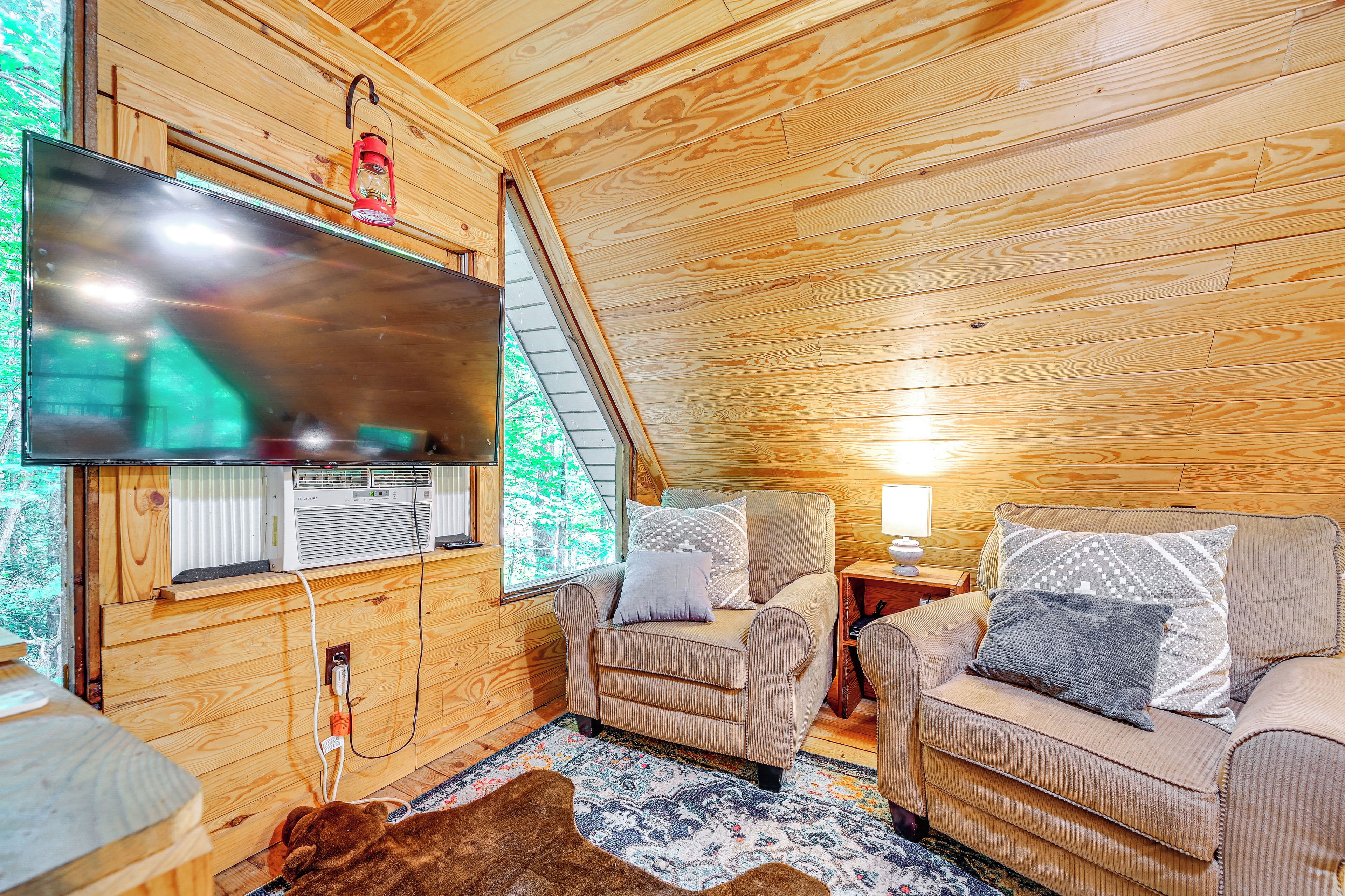 A cozy cabin interior with a large TV, two armchairs, a wooden ceiling, and a window overlooking greenery.
