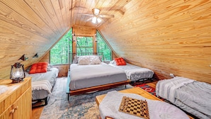 A cozy attic bedroom with wooden walls, a sloped ceiling, and a large window offering a view of the forest. There are two beds, a wooden dresser, and a chessboard on the floor. - Cozy Nature Escape! Roaring Gap Cabin w/ Patio (Roaring Gap)