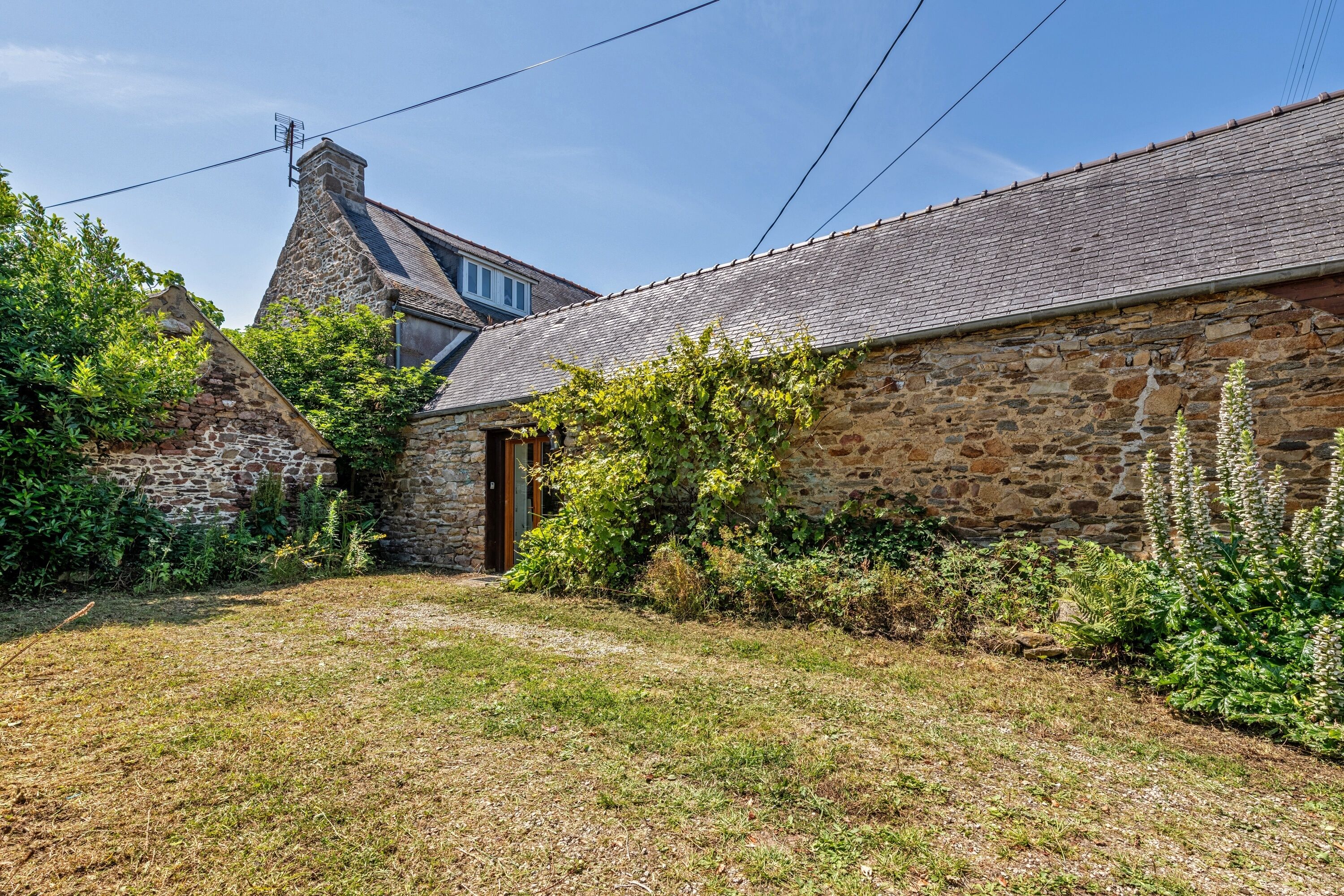 A stone building with a sloped roof, surrounded by greenery and a clear sky.