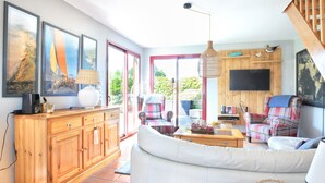 A spacious living room with a large sofa, a wooden cabinet, and a television mounted on a wooden wall. - La Maison Merlin - Maison avec piscine, Sénoville (Sénoville)