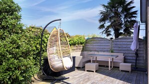 A patio with a hanging chair, wicker furniture, and a wooden deck. - La Maison Merlin - Maison avec piscine, Sénoville (Sénoville)