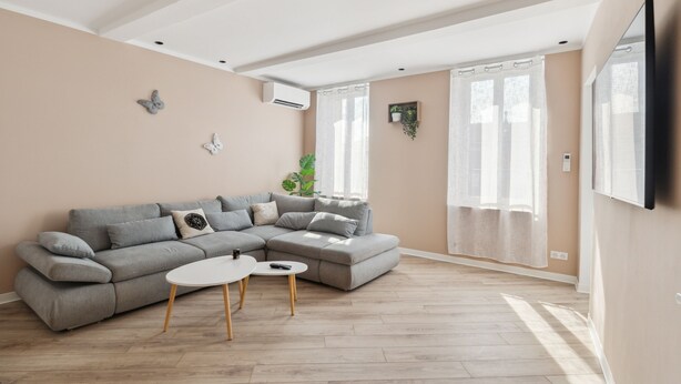 Un salon moderne avec un canapé d’angle gris, une petite table basse blanche et une grande fenêtre ornée de rideaux légers.
