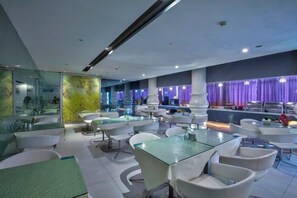 A modern hotel lobby with glass tables, white chairs, and a reception area with purple lighting.