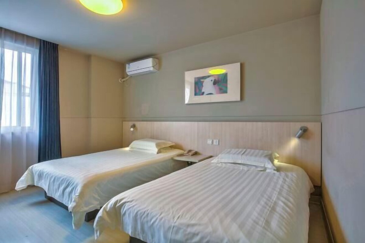A hotel room with two single beds, a wall-mounted air conditioner, a framed picture, and a bedside table with a lamp.