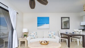 Ein modernes Wohnzimmer mit einem runden Holztisch, einem weißen Sofa mit Kissen mit Sternenmuster, einem Esstisch mit Stühlen und einem Gemälde einer Strandansicht an der Wand.