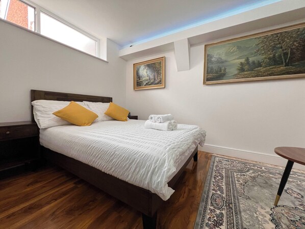 A neatly arranged bedroom with a wooden bed, white linens, and two yellow pillows. There are two paintings on the wall, a small table with a chair, and a patterned rug on the floor.