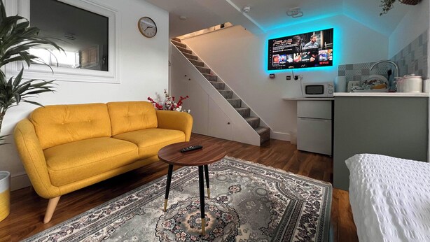 A modern living space with a yellow sofa, a patterned rug, a staircase, a kitchen area, and a wall-mounted TV displaying a streaming service. - Central Luton Executive Suite (Luton)