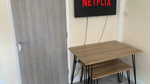 Huoneessa on puinen ovi, pöytä ja penkki sekä seinälle asennettu televisio, jonka näytöllä näkyy teksti ”NETFLIX”.