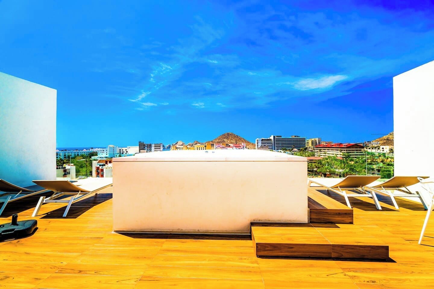 Un bar en la azotea con una amplia mesa blanca y sillones, con vistas a un paisaje urbano que incluye edificios y una montaña.
