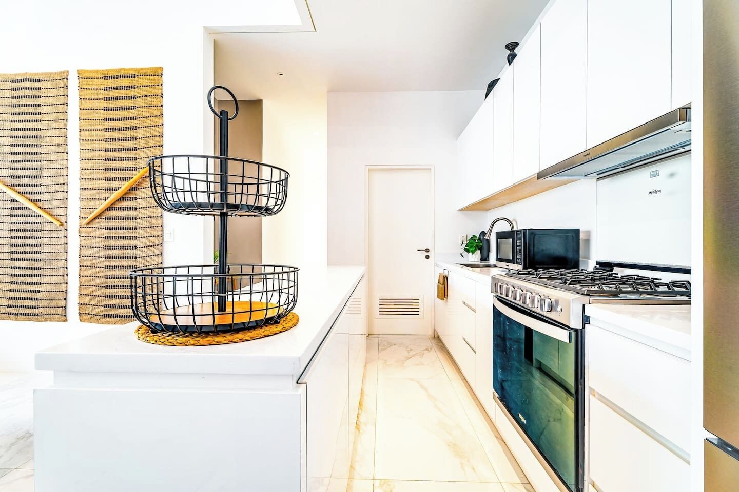 A modern kitchen with white cabinets, a double oven, and a microwave. There is a black wire basket on a white countertop.