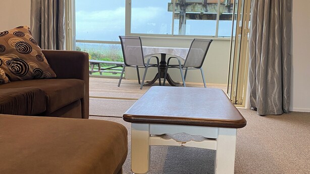 Ein Zimmer mit einem braunen Sofa, einem weißen Tisch und Blick ins Freie durch große Fenster.