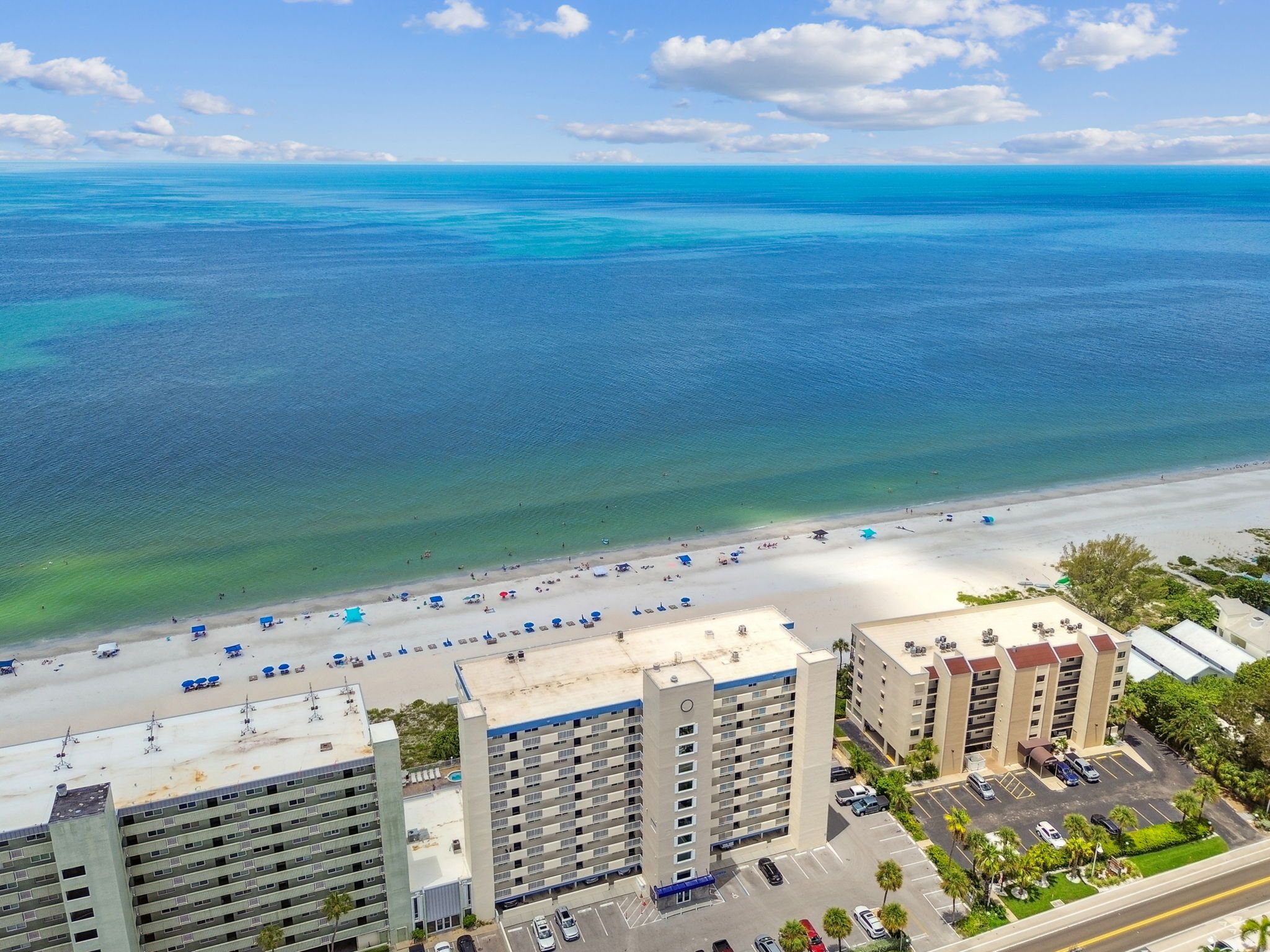 A beachfront hotel with multiple floors, a parking area, and a beach with umbrellas and people.