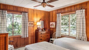 Habitación con paredes de madera, dos camas, ventilador de techo, cómoda, silla y ventanas amplias con cortinas a cuadros.
