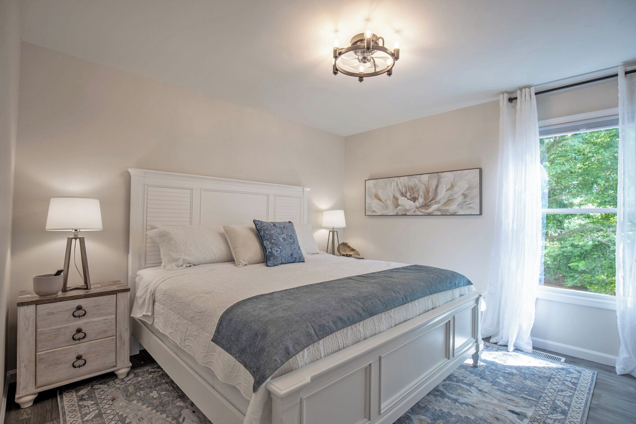 A bedroom with a large bed, bedside lamps, a nightstand, a window with curtains, and a wall-mounted artwork.