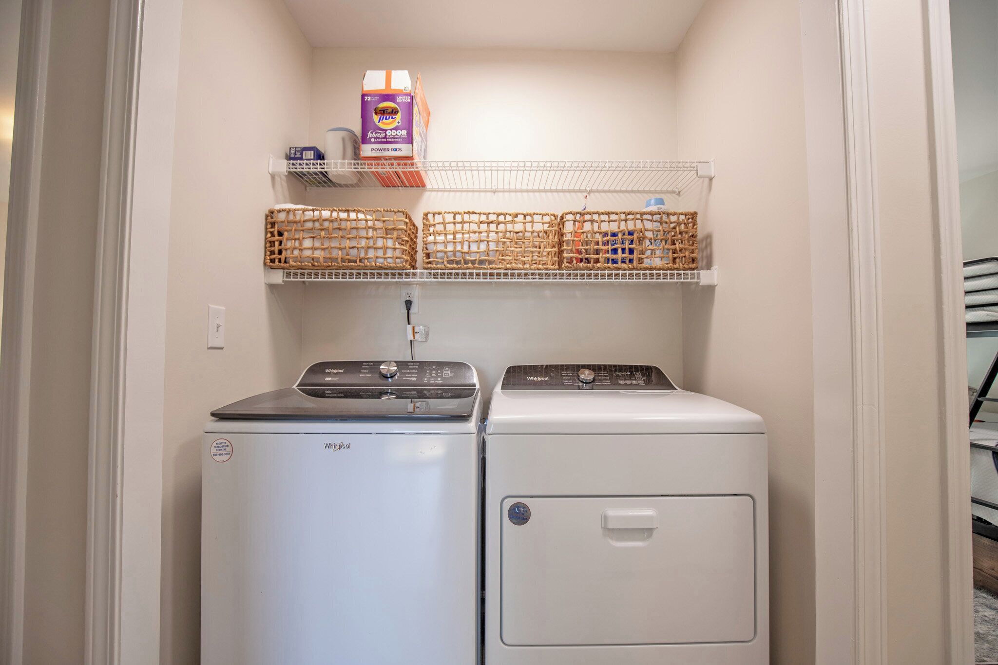Une buanderie équipée de deux machines à laver et de deux sèche-linge à chargement frontal, d’une étagère avec des paniers et d’un carton de lessive.