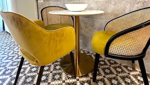 A modern dining area with a round table, two yellow chairs, a white bowl, a flat-screen TV mounted on a stone wall, and a patterned floor. - Balcony Hotel (St. Julian's)
