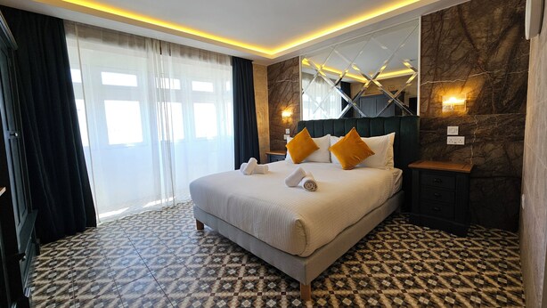 A hotel room with a large bed, a nightstand, a mirror, and a patterned floor. - Balcony Hotel (St. Julian's)