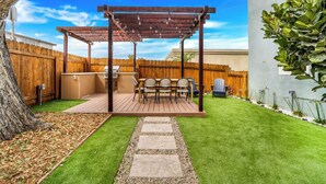Un patio trasero con una terraza de madera, una pérgola con luces colgantes, una zona de barbacoa y un camino de piedra.