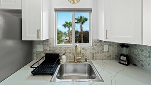 Una cocina con armarios blancos, un lavamanos doble, un ventanal con vista a palmeras y un salpicadero de azulejos hexagonales.