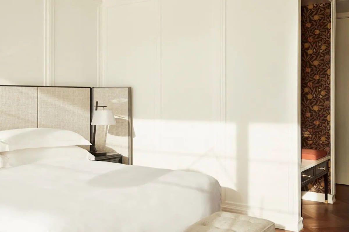Una cama bien hecha con sábanas blancas, una mesita de noche con lámpara y una pared con papel tapiz estampado.