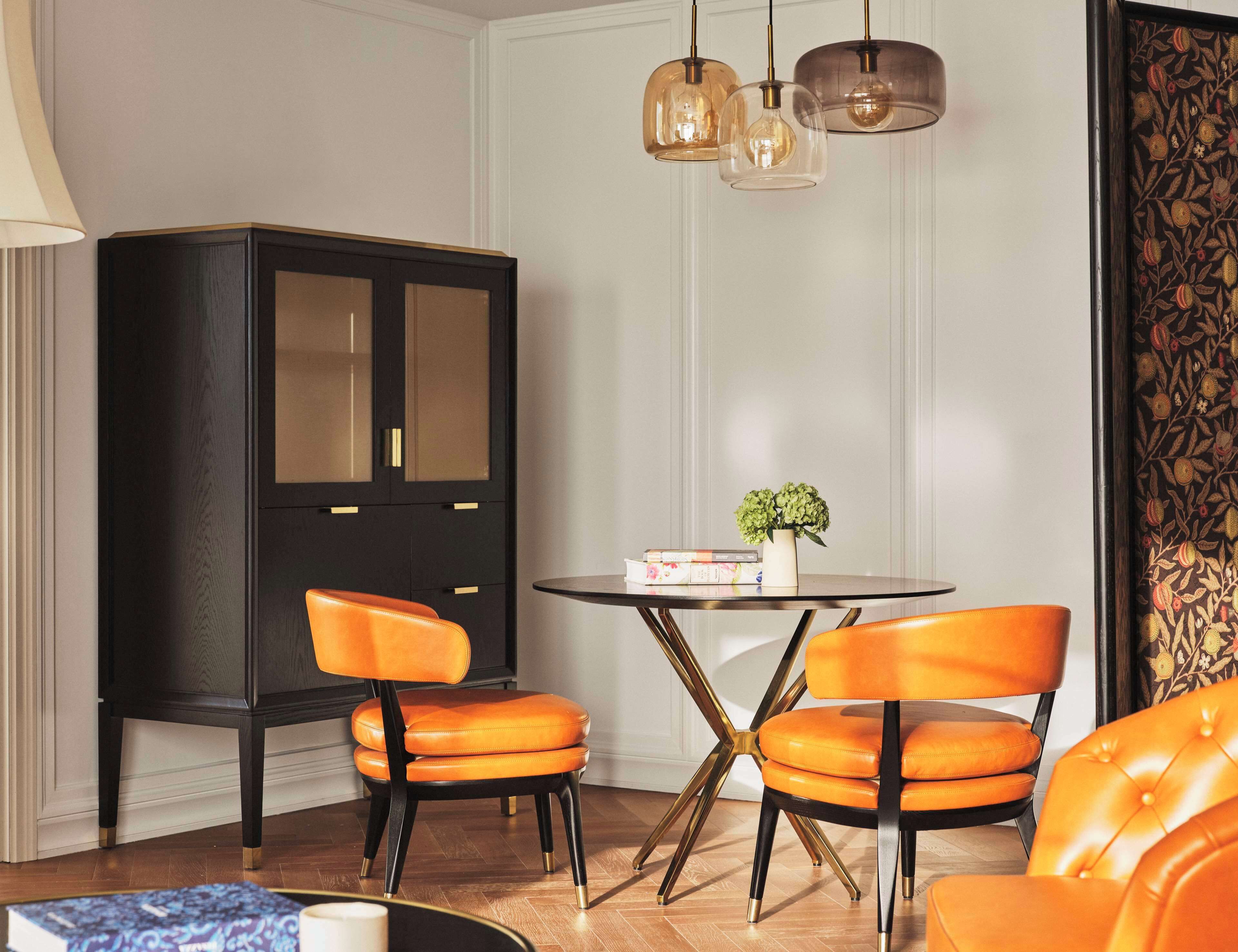 A dining area with a black cabinet, two orange chairs, a round table, and a vase with green flowers.