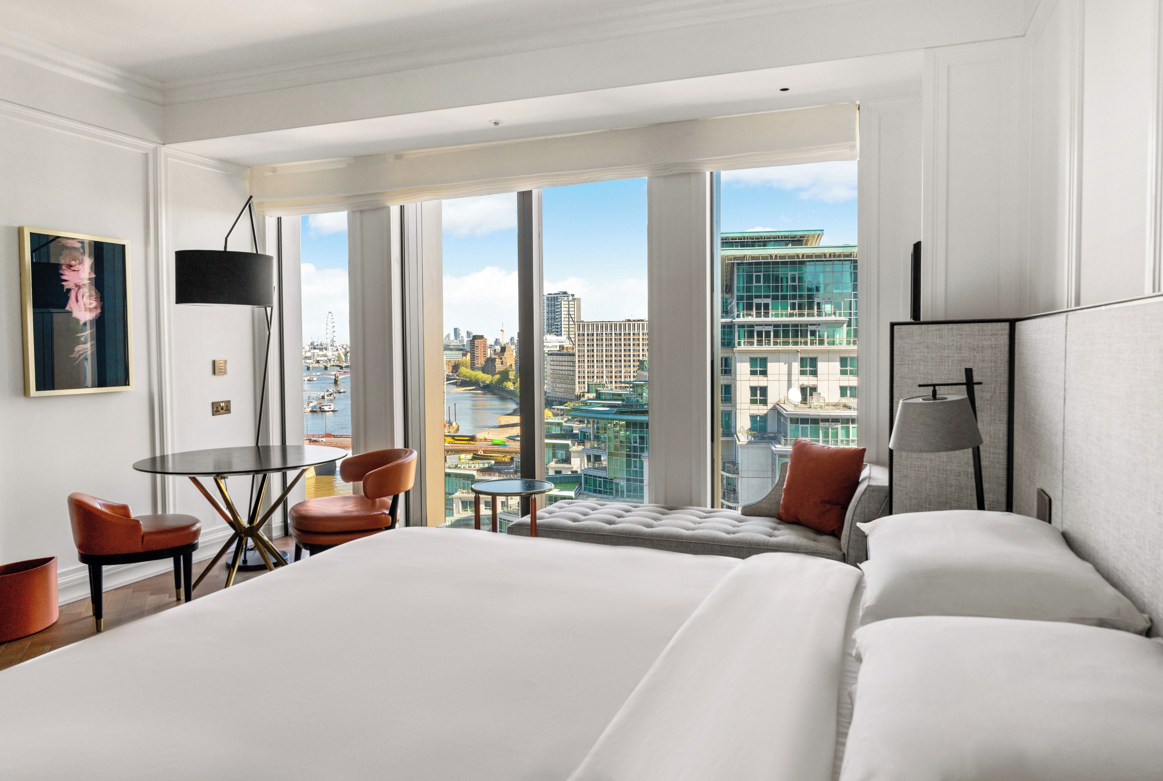 Habitación de hotel con una cama grande, una mesita, dos sillas, un sofá y vistas a la ciudad a través de amplios ventanales.