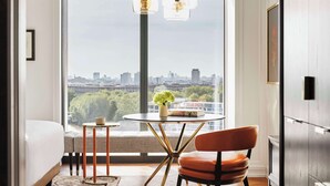 Een moderne slaapkamer met een groot raam, een rond glazen tafeltje met een boek en een vaas, een bruine leren fauteuil, en een bed met een wit hoofdeinde.