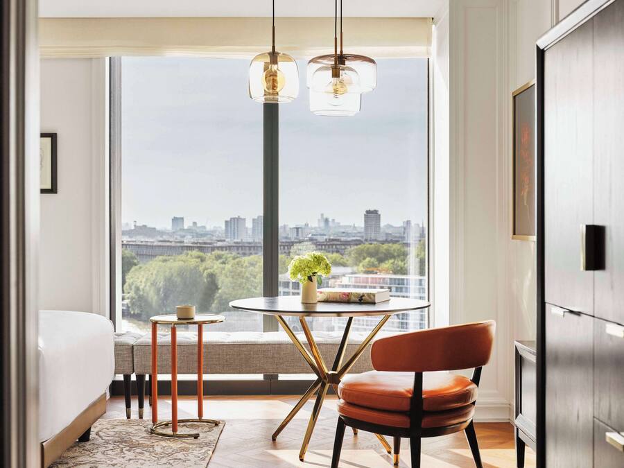 Ein modernes Schlafzimmer mit großem Fenster, einem runden Glastisch mit Buch und Vase, einem braunen Ledersessel und einem Bett mit weißem Kopfteil.