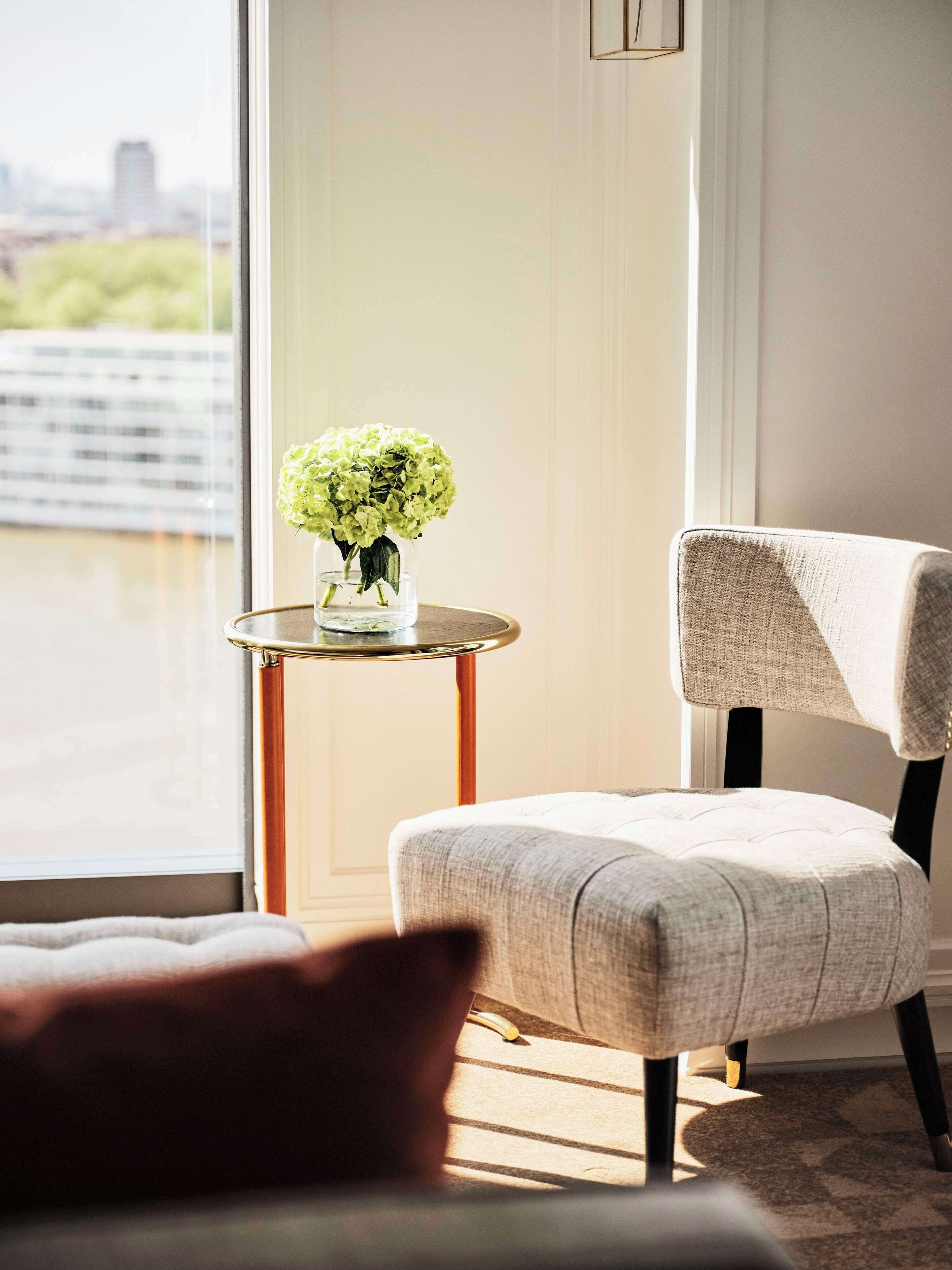 A room with a view of a cityscape through a large window, a small round table with a vase of flowers, and a chair with a high backrest.
