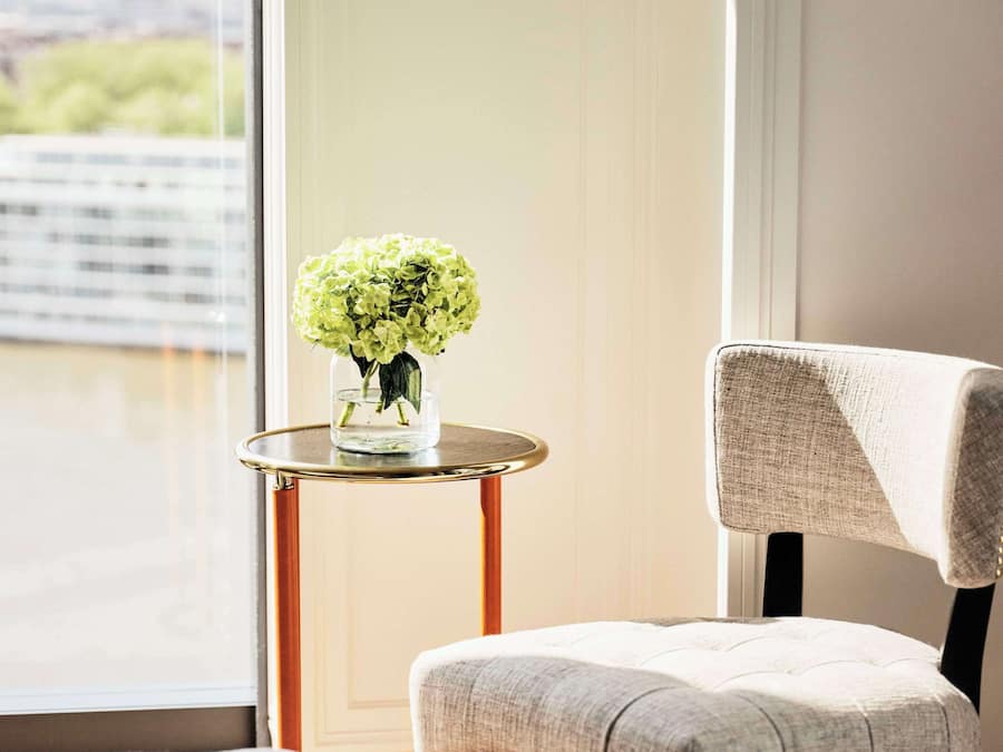 Una habitación con vistas a un paisaje urbano a través de un ventanal grande, una mesita redonda con un florero y una silla con respaldo alto.